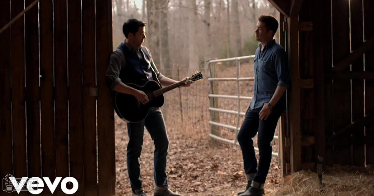 Two young adult males standing in a rustic barn setting with wooden walls, metal gate, hay on the ground, and bare trees suggesting autumn season; one wearing a dark blue shirt and jeans, the other in a dark blue button-down and bracelet.