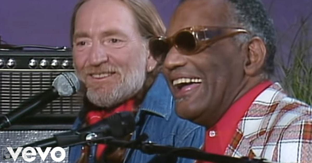 Middle-aged to older adult male singer wearing a blue denim jacket with a red plaid shirt, performing on stage with another older adult male singer in a red and white plaid shirt; purple backdrop suggests an outdoor or stage setting.