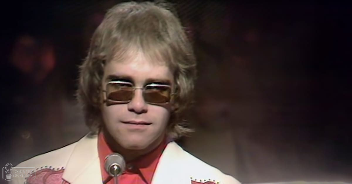 Young adult male singer in a white suit with red accents performing on stage, spotlight lighting creating dramatic shadows.