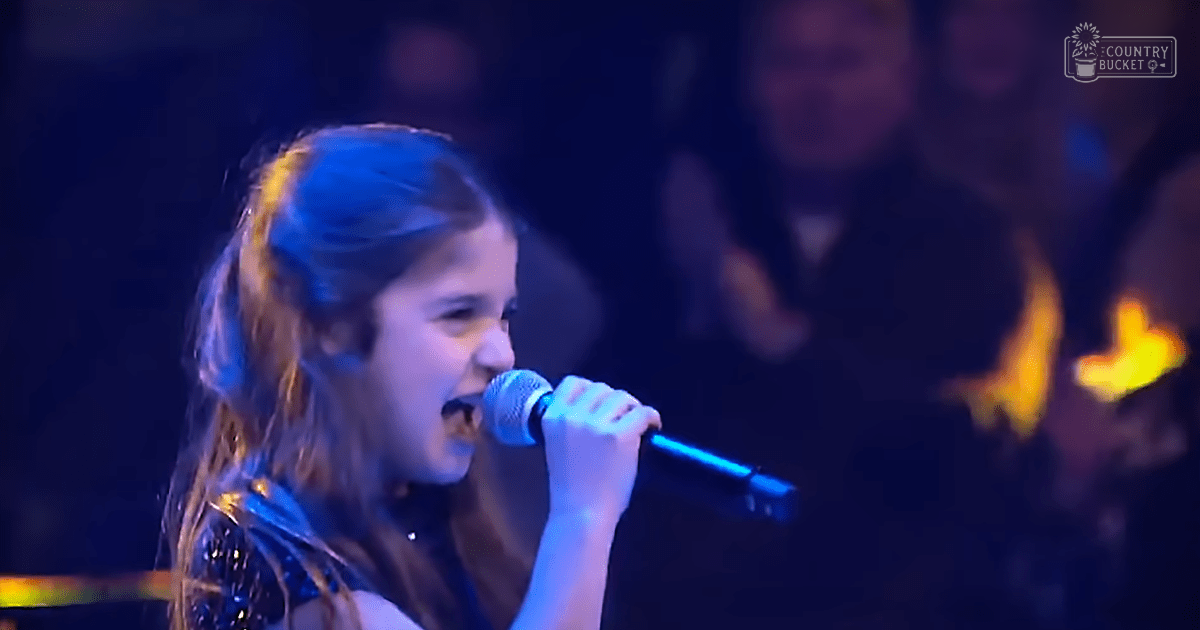 A young adult female singer performing on a stage with a sparkly dark blue dress and reflective fabric, holding a microphone under stage lighting at an indoor concert.