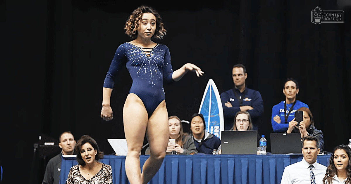 A young adult female gymnast competes indoors, surrounded by judges and spectators dressed in casual to semi-formal attire under artificial lighting.
