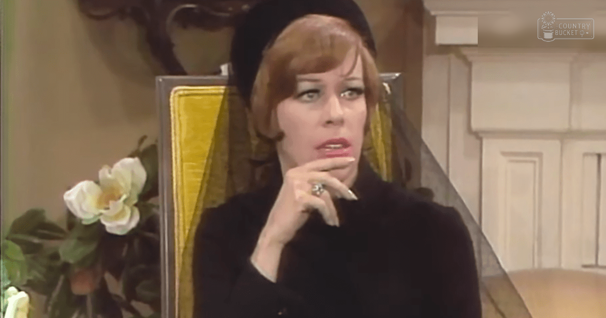 An adult female wearing a black fitted garment with long sleeves and a high neckline, standing indoors near a yellow upholstered chair and white floral arrangement.