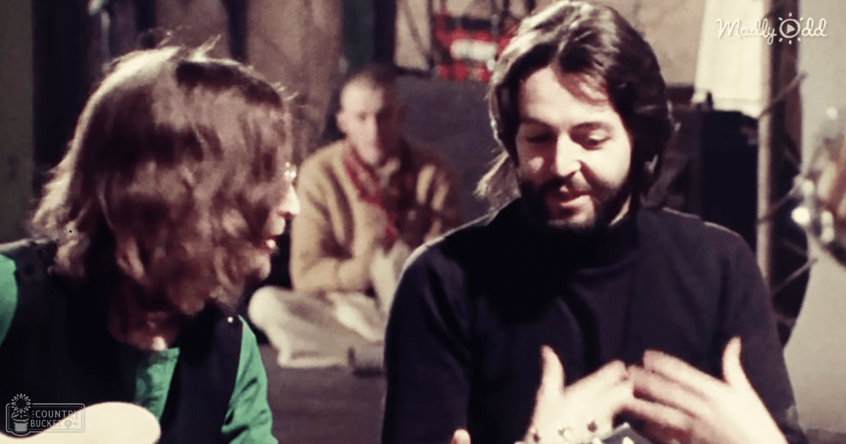 A man with long hair and a beard in an indoor studio or recording room, wearing a black turtleneck sweater, surrounded by another adult female in a green top under a black vest and an adult male in the background wearing a beige jacket and red scarf.