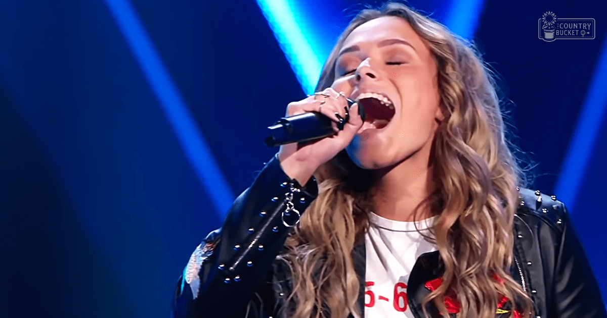 A young adult female in a black leather jacket and white T-shirt with '5-6' printed on it, performing on stage under blue stage lights.