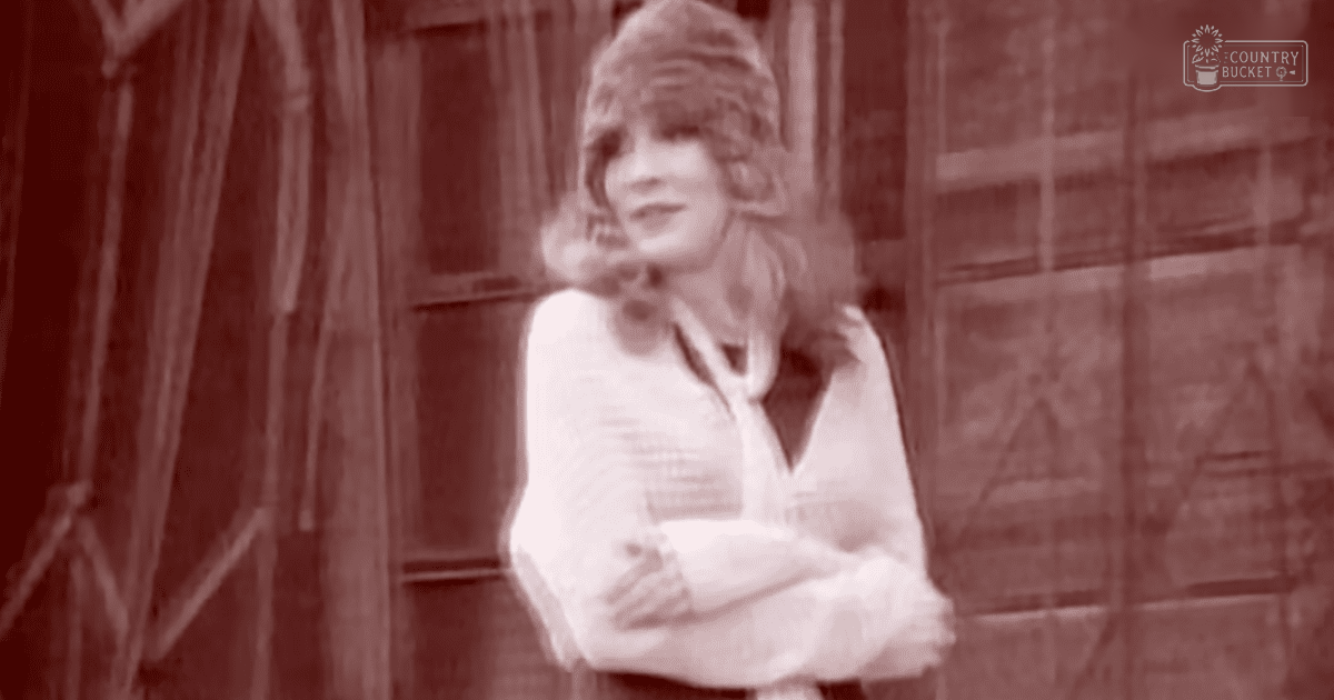 A young adult female wearing a loose, flowing white V-neck blouse stands against an urban backdrop with a wooden facade.