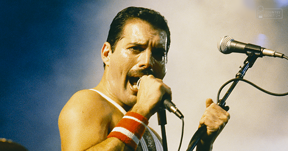 An adult male singer on stage wearing a white tank top with red and white striped wristbands, holding a microphone, performing at what appears to be a concert.