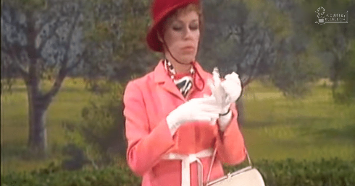 A woman wearing a bright pink coat with long sleeves, white gloves, and a red hat stands outdoors amidst greenery. The scene suggests spring or summer day.