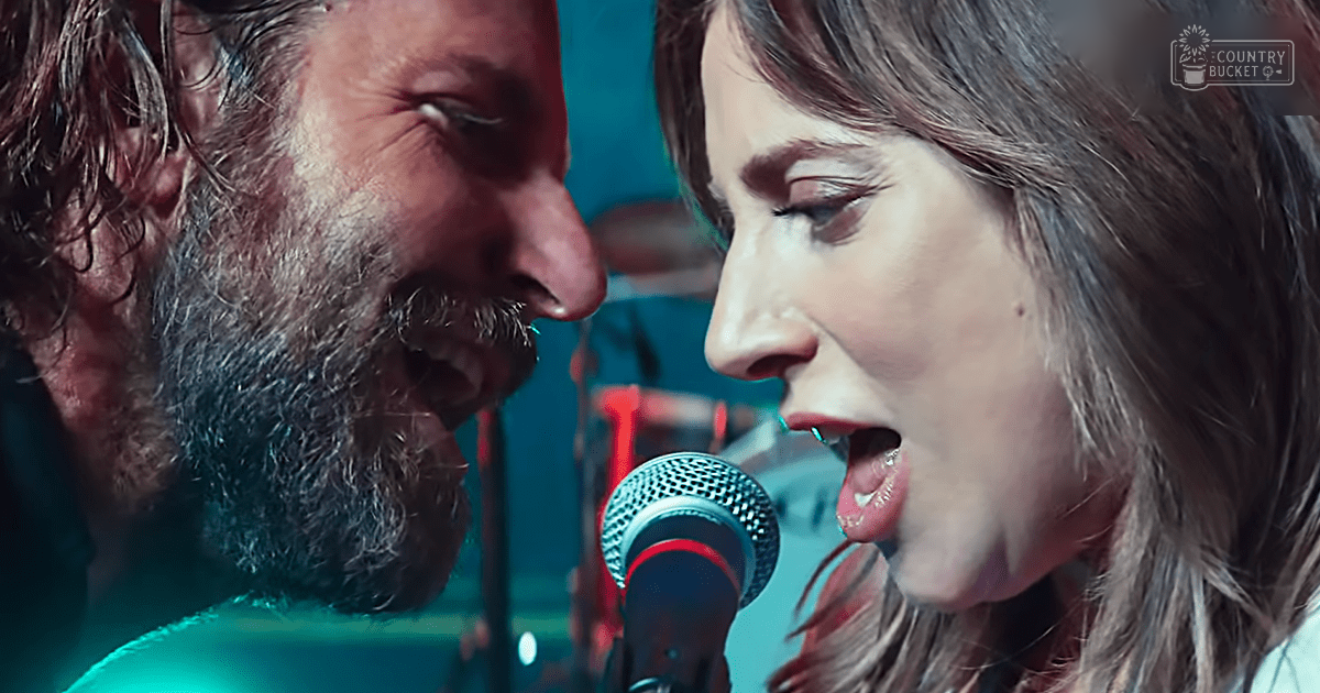 Middle-aged male singer in dark shirt performing next to young adult female with light top, microphone stand and blue background suggest an indoor musical performance setting.