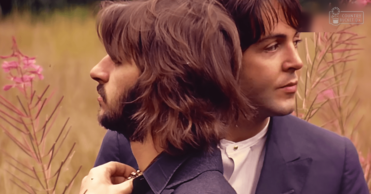 Two young adult males in tailored blazers from the 1960s, wearing white shirts and a watch, standing outdoors with pink flowers and greenery.