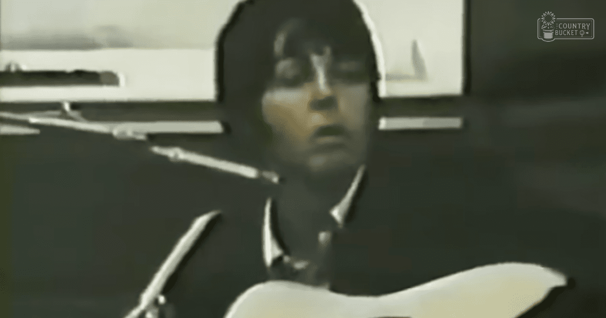 An adult male wearing a white collared shirt with short sleeves stands indoors, possibly in a recording or performance space, with even artificial lighting and neutral tones.