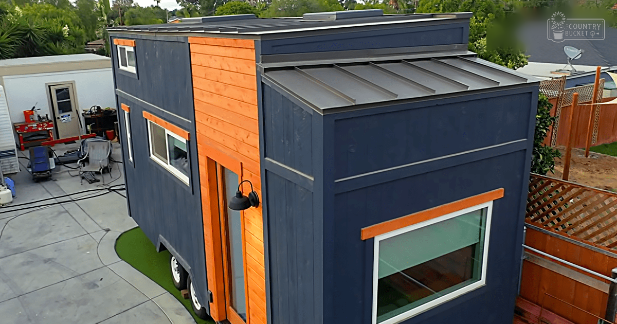 A tiny house with dark blue and orange paneling situated on a small patio area surrounded by lush greenery in an outdoor residential setting during spring or summer.