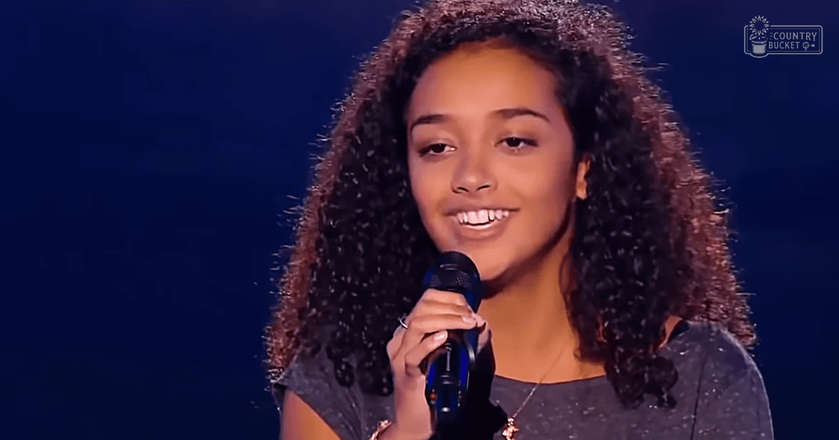 A young adult female wearing a dark gray or black, speckled short-sleeved top with a scoop neckline and relaxed fit, accessorized with a necklace, performing on an indoor stage with 'Madly Odd' text visible in the background.