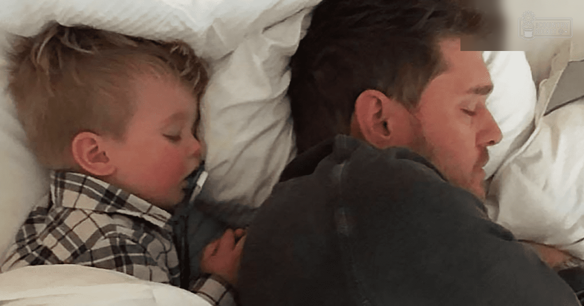 A toddler boy wearing a plaid shirt sleeps next to an adult male in a softly lit bedroom at night.