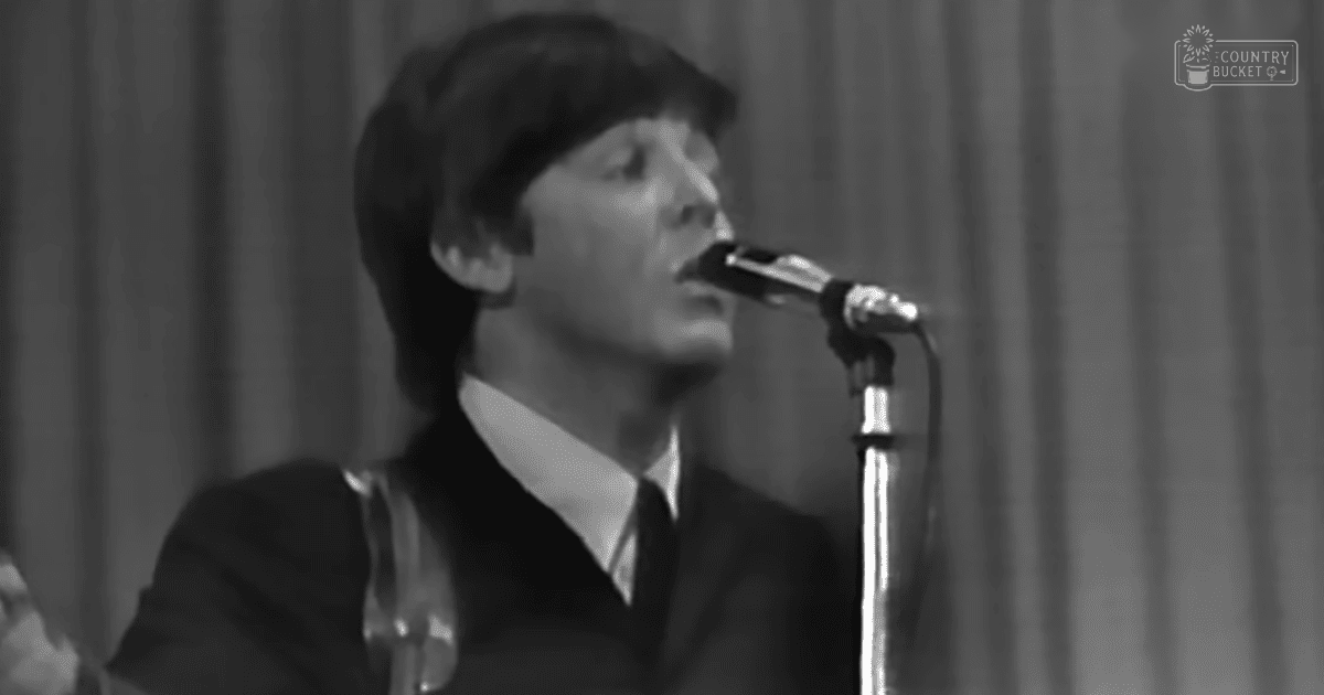 A young adult male singer performing at an indoor event wearing a dark suit jacket with a white collared shirt and tie, holding a guitar strap over his right shoulder.