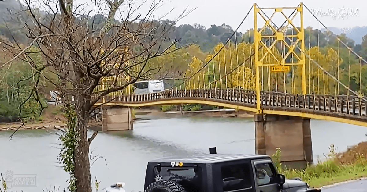 A serene yellow suspension bridge spans a calm river, surrounded by trees and hills under an overcast sky in autumn.