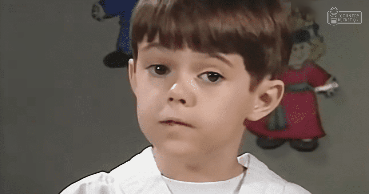 A young male child wearing a white historical-style garment indoors with two painted figures on the wall, neutral-colored walls, muted tones.