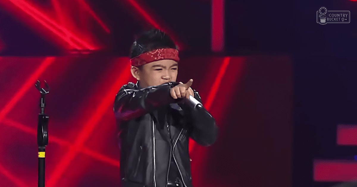 A young male child in a black leather jacket and red bandana performs on stage under dramatic red lighting.