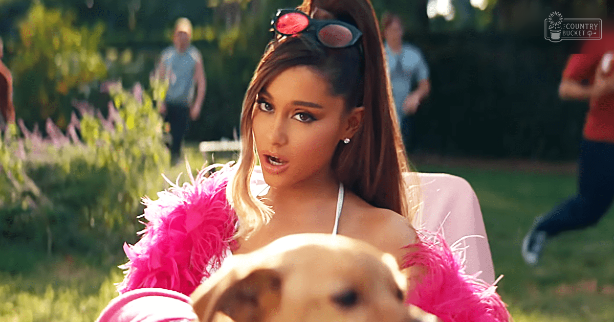 A young adult female wearing a white spaghetti strap top and pink feather boa stands outdoors, surrounded by adults and a golden retriever dog. The scene is set in what appears to be a park or garden during spring or summer with bright daylight.