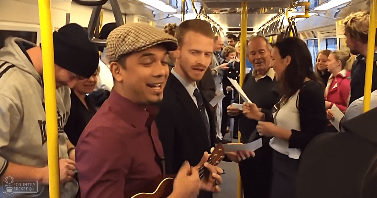 A young adult male playing a ukulele on a public transportation bus with yellow poles and hanging straps, surrounded by other passengers in various attire.