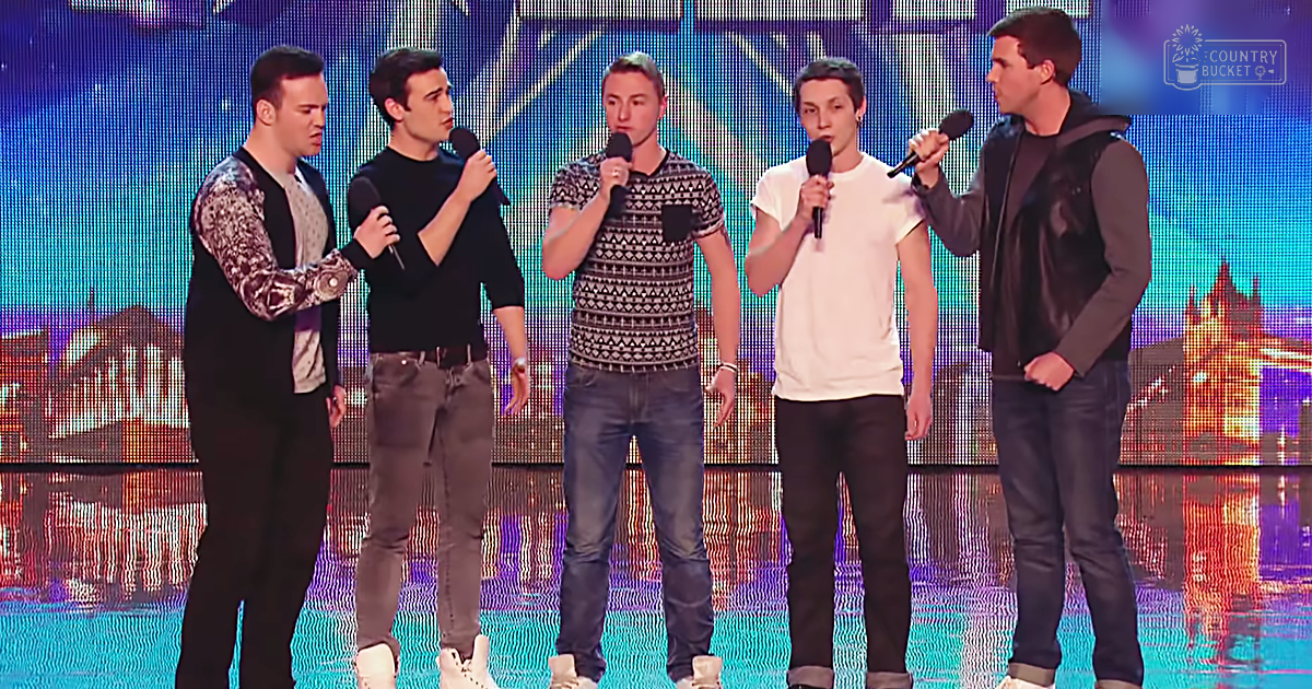 A group performance scene with young adult males in patterned sweaters and V-necks against a backdrop of an illuminated LED screen displaying London's landmarks at night, set indoors under artificial evening lighting.