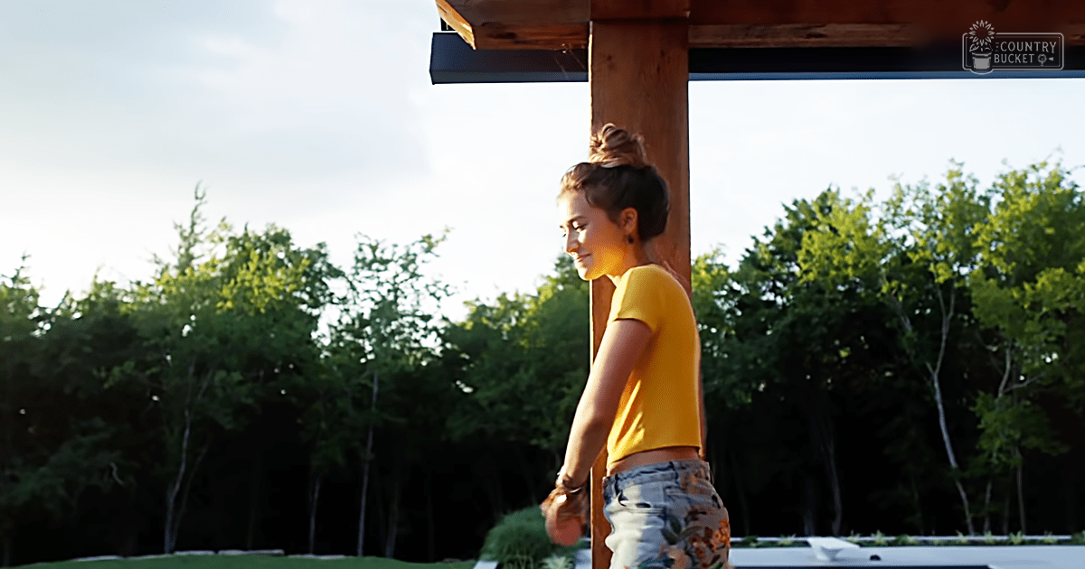 A young adult female wearing a yellow crop top and light blue jeans with camouflage on the lower legs, standing outdoors amidst trees under clear daylight in spring or summer.