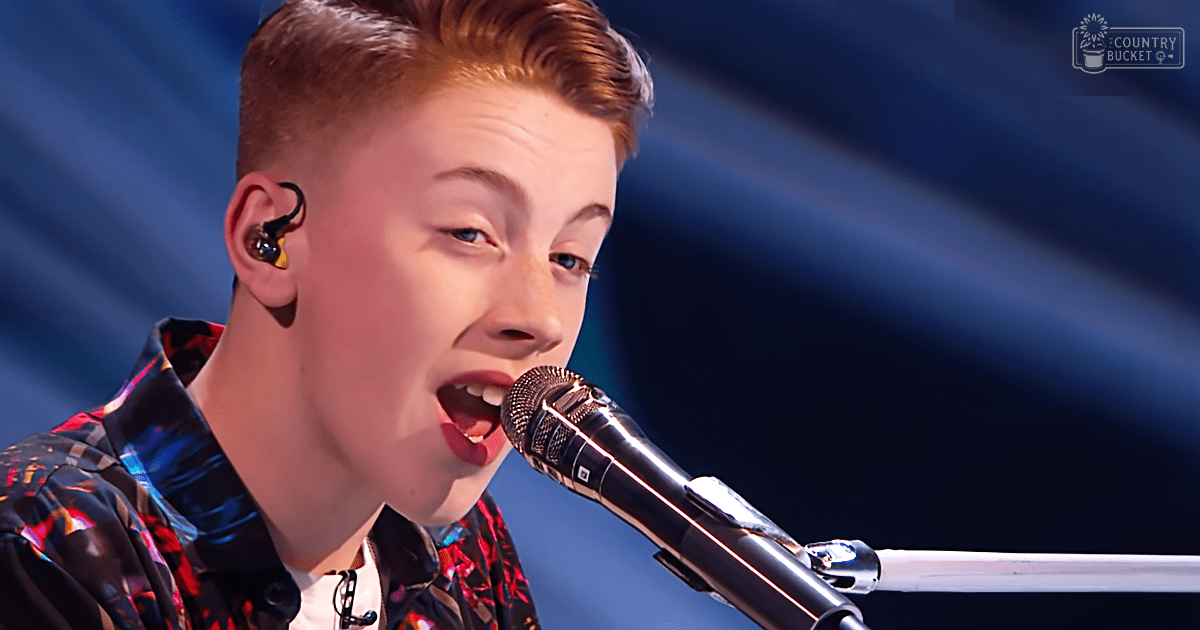 Young adult male in a patterned button-down shirt performing on an indoor stage with dark blue curtains and lighting effects suggesting evening or night time.