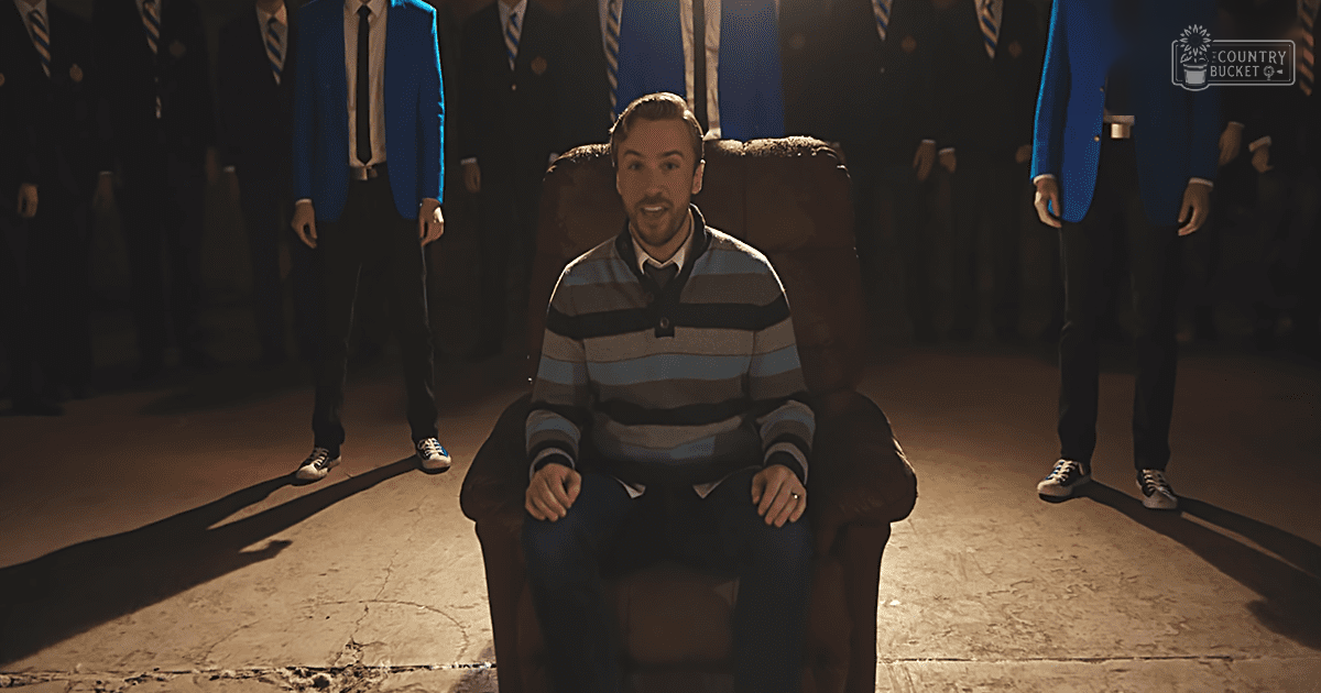 A young adult male sits in a brown armchair indoors, surrounded by adults dressed in formal attire. The scene is dimly lit with shadows cast by the standing individuals, suggesting an evening setting.