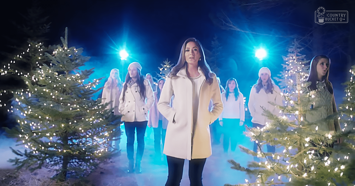 A young adult female in a cream coat stands outdoors with Christmas trees, surrounded by other individuals dressed for winter.