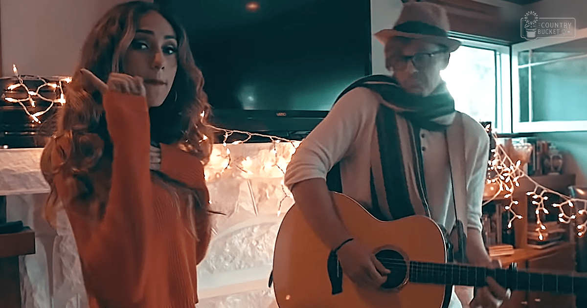 Adult female in orange ribbed sweater and adult male holding acoustic guitar during musical performance, cozy indoor setting with string lights and natural light.