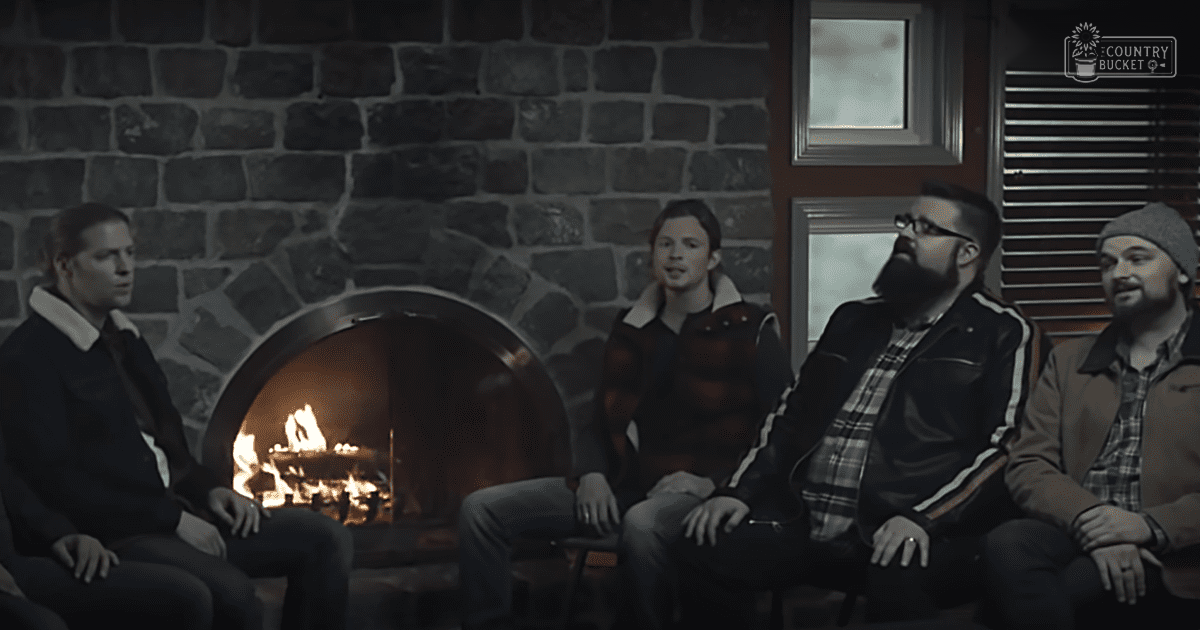 A group of adults gathered indoors in front of a cozy stone fireplace with fire burning inside it, wearing jackets and pants, under warm ambient light suggesting evening or nighttime.
