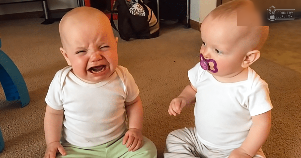 Two infants wearing white short-sleeved onesies, one with a light green pant and another possibly male or gender-neutral. Setting appears to be an indoor carpeted home space.