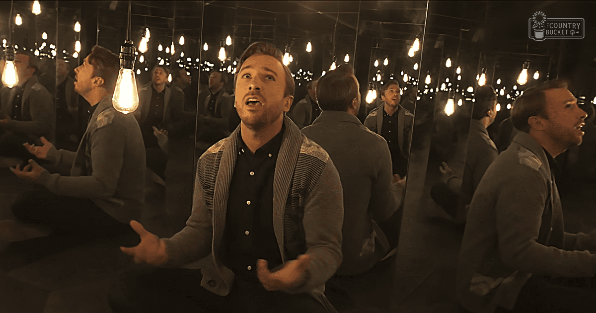 A young adult male in a dimly lit room filled with hanging vintage-style filament lights and multiple mirrors reflecting his image.