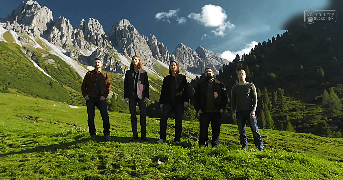 Five adults posing outdoors against a backdrop of rugged mountains under a clear blue sky with scattered clouds.