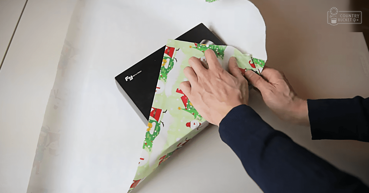 An adult male wrapping a gift with holiday-themed wrapping paper, indoors in natural light.