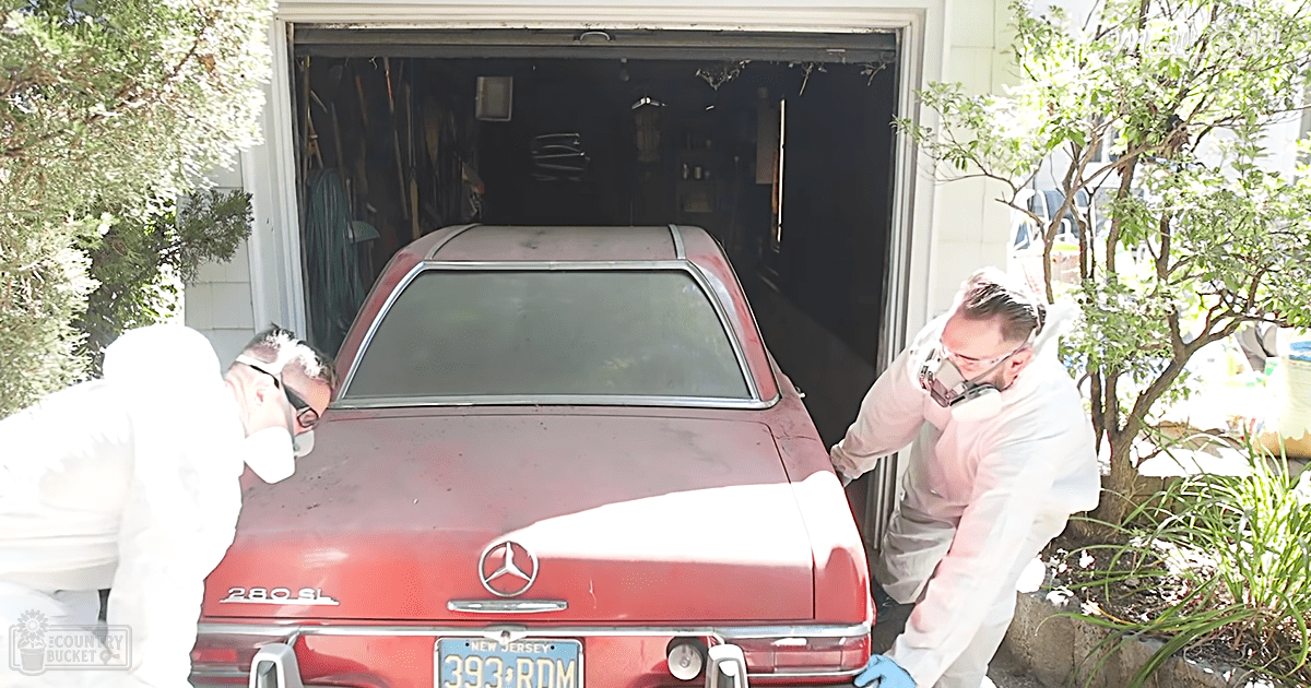 Two adult males wearing white coveralls with hoods and gloves, working on a red Mercedes-Benz 280SL parked outside an open garage door in front of a residential home. The car's license plate reads '393 RDM'.