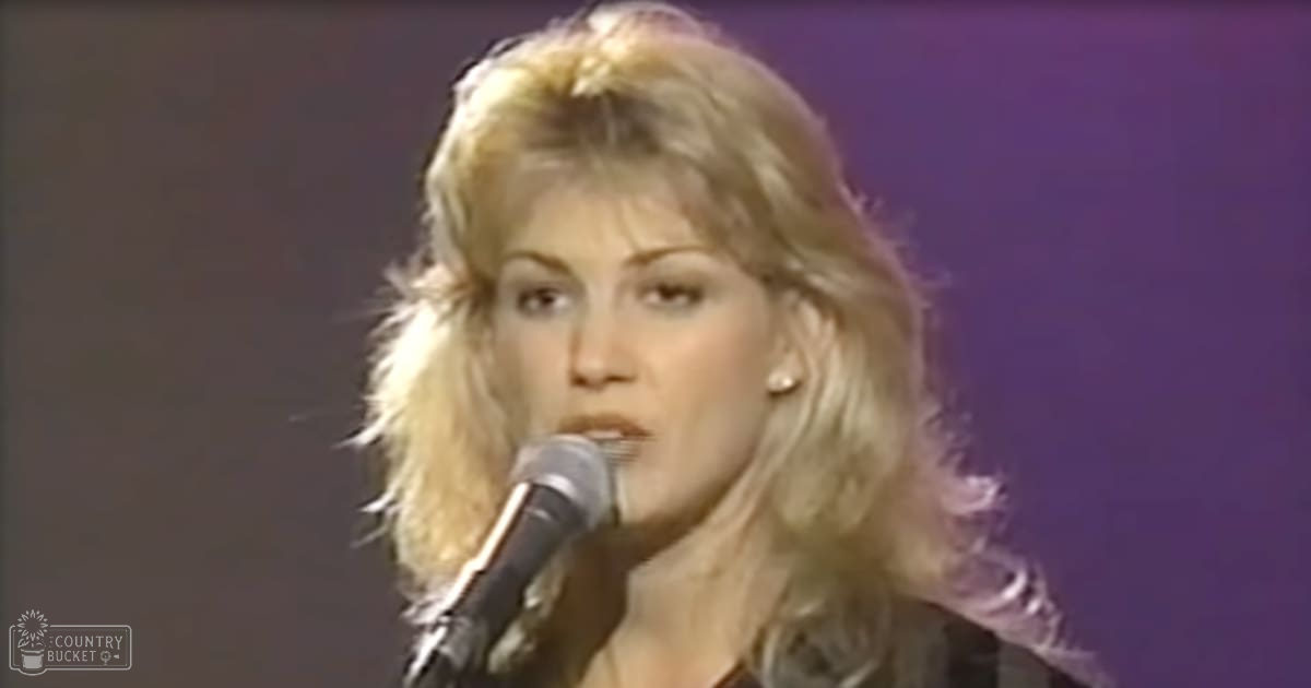 An adult female singer wearing a dark, long-sleeved top with high neckline and small earrings, performing into a microphone under artificial purple stage lighting.