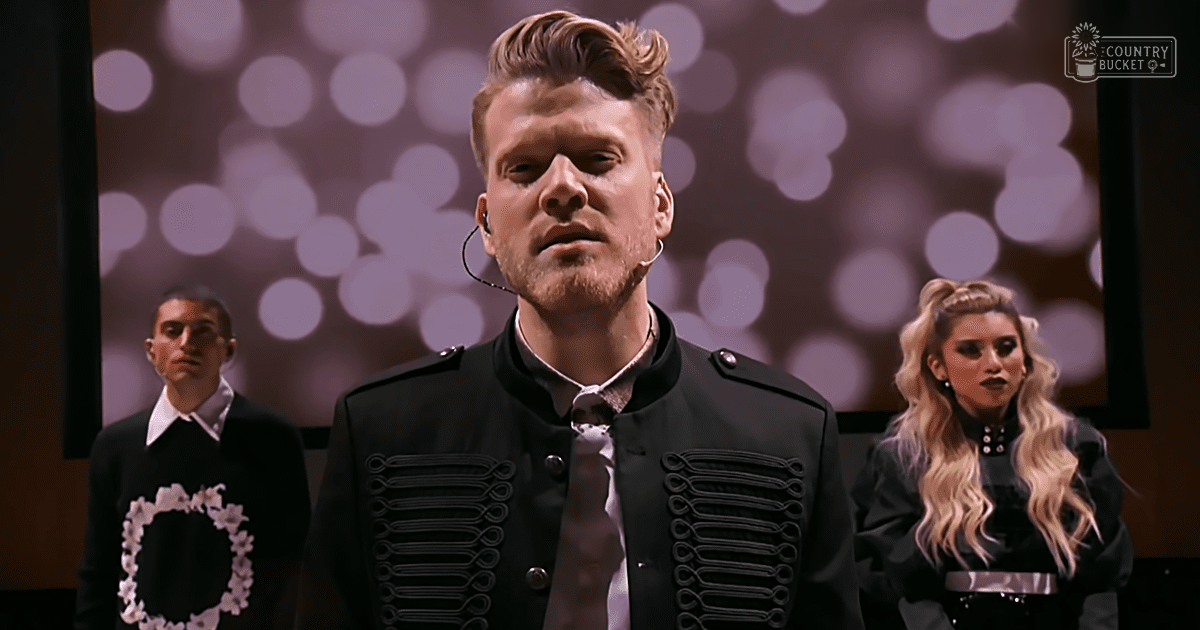 Young adult male in a black military-style jacket with silver buttons over white shirt and tie, earpiece microphone; teen or young adult male in dark sweater with floral wreath design on stage backdrop with bokeh lights.