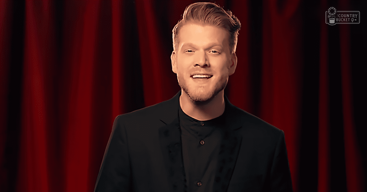 A young adult male in a dark-colored suit jacket and shirt, standing against red curtains indoors.