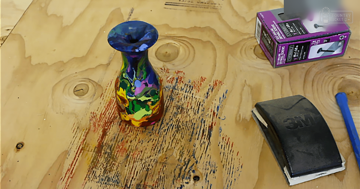 A colorful glass vase with abstract patterns sits atop a wooden surface indoors, surrounded by tools like screws and sanding blocks.