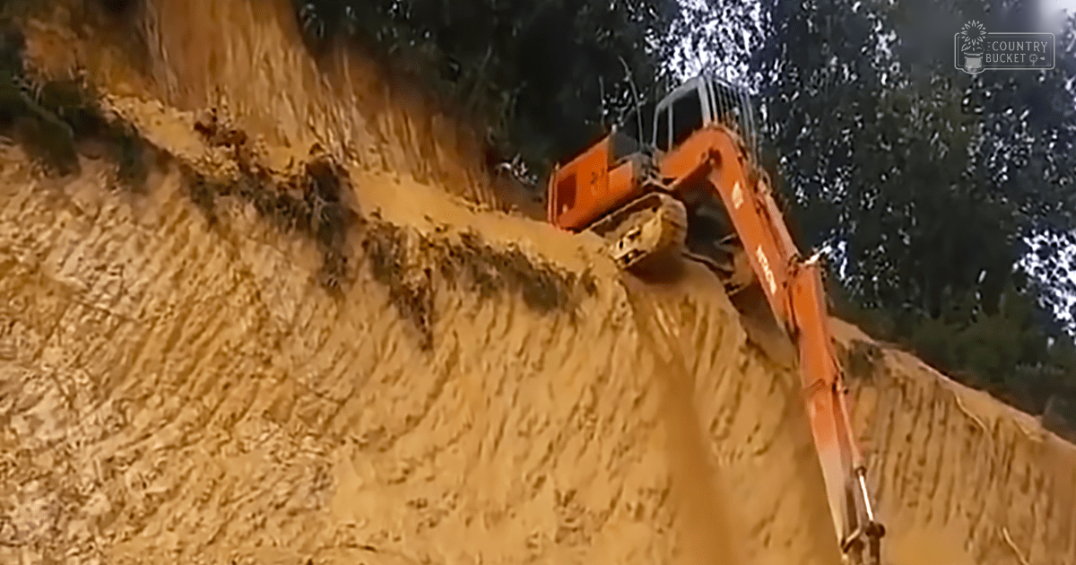 An excavator actively digging in a sandy area at a construction site with trees in the background, suggesting spring or summer.