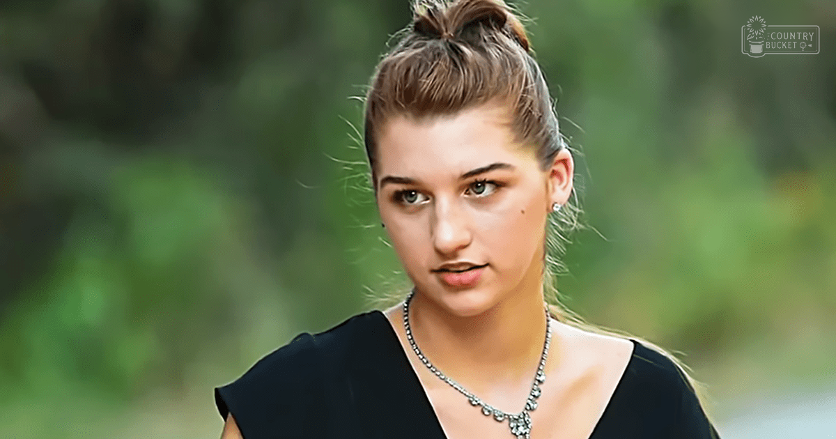 A young adult female wearing a black sleeveless top and small earrings, surrounded by lush greenery under bright natural light.