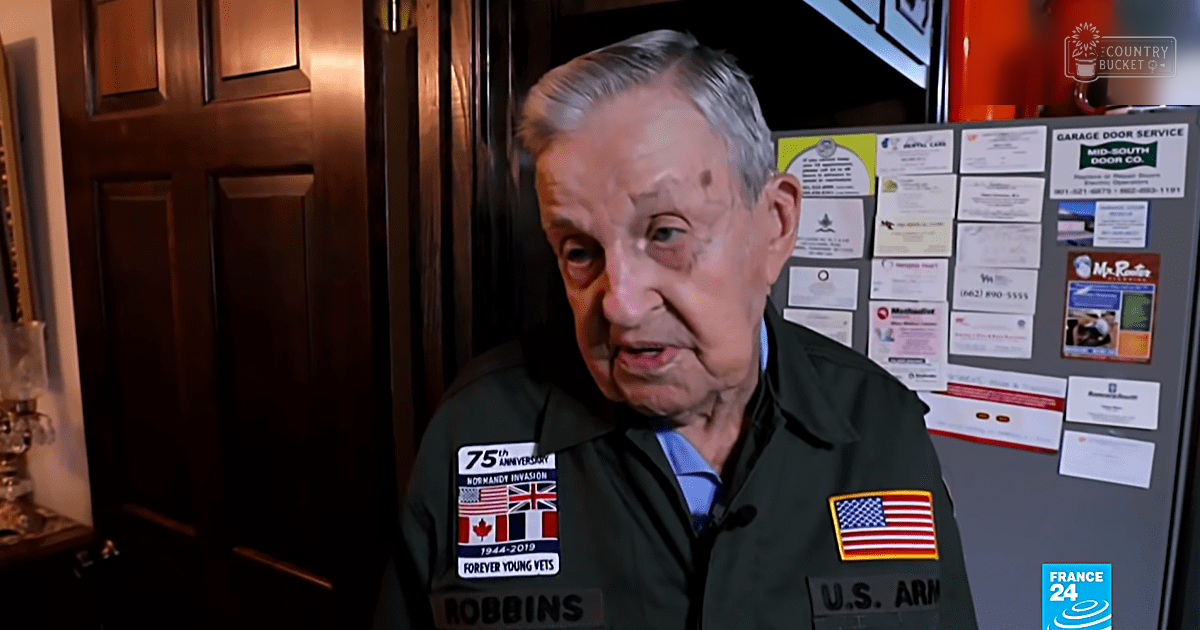 An older adult male wearing a dark green U.S. Army jacket with commemorative patches, indoors in a home setting.