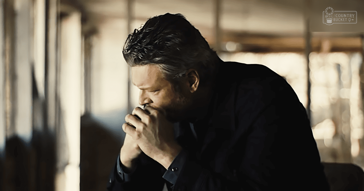 A middle-aged adult male in a relaxed fit dark-colored jacket and collared shirt stands indoors near large windows allowing natural light, suggesting daytime during cooler weather.
