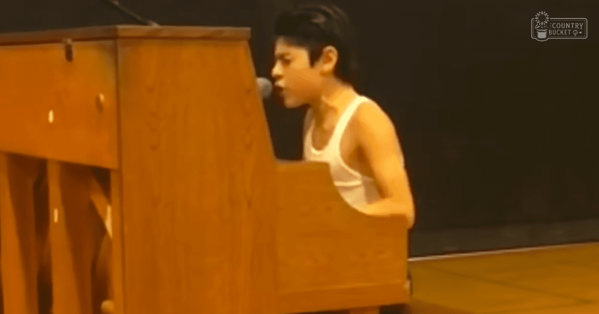 A young adult male in a white tank top and black cap stands on an indoor stage with a wooden podium and microphone, suggesting a formal event like a concert or speech. The setting is dimly lit with brown tones from the podium and white from his attire.