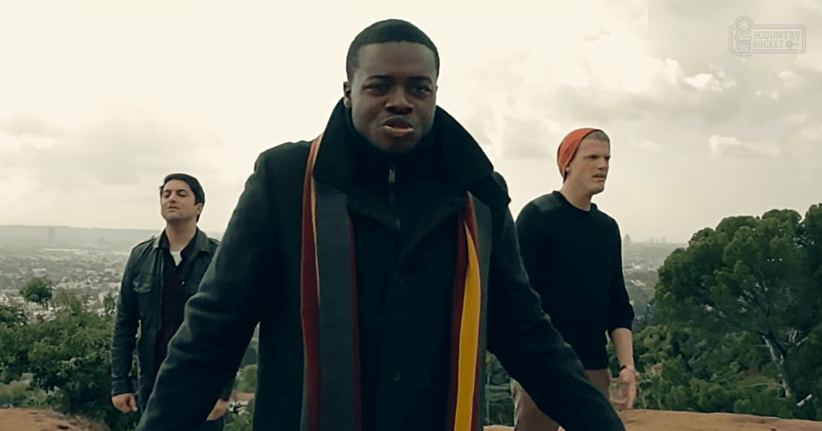 Young adult male in a black coat with red/yellow stripes on sleeves and an orange headband, standing outdoors against an urban backdrop under cloudy skies.