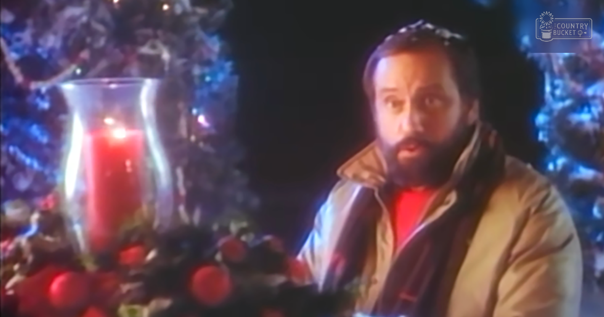 A middle-aged male in an indoor holiday setting, wearing a beige jacket and red garment with dark scarf, surrounded by Christmas decorations.