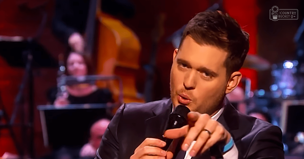 A young adult male singer in a dark suit jacket with white shirt, holding a microphone and wearing a ring, performing on stage in an indoor concert hall setting.