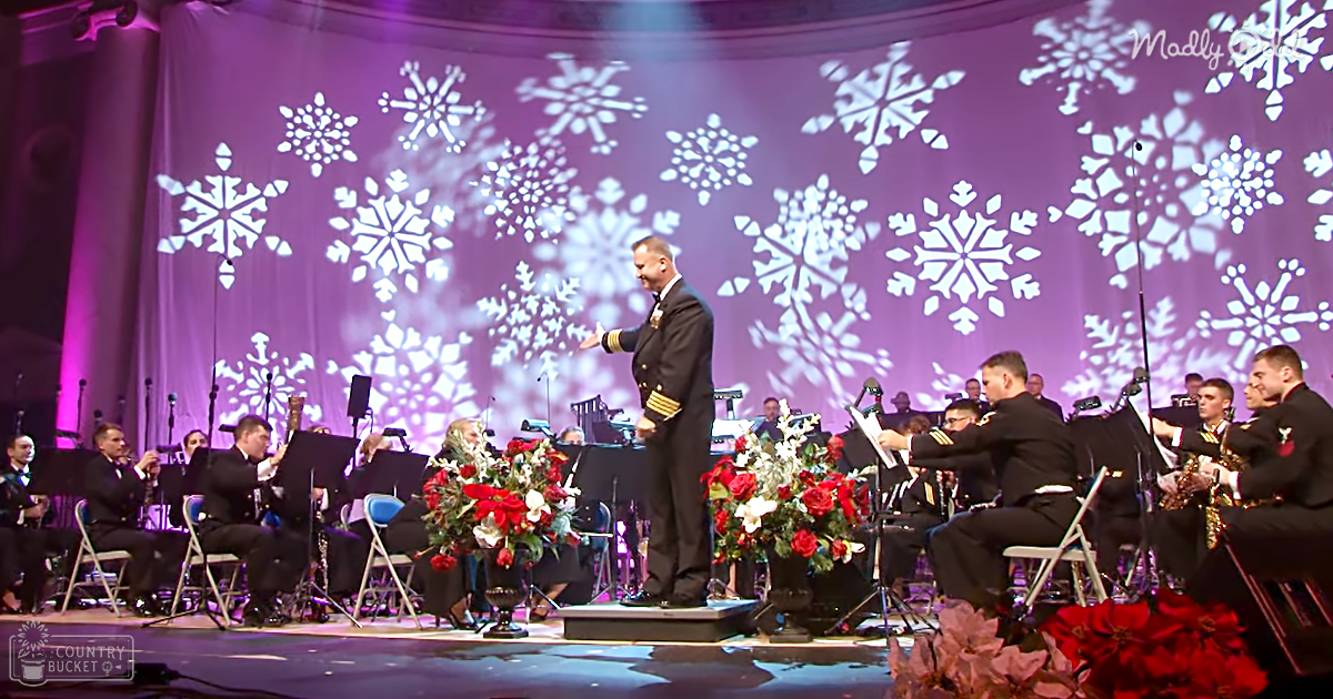 Conductor leading an orchestra in a formal event at what appears to be a concert hall with adult males in their 30s-50s dressed in black uniforms, surrounded by floral arrangements and snowflake patterns projected on a purple backdrop.