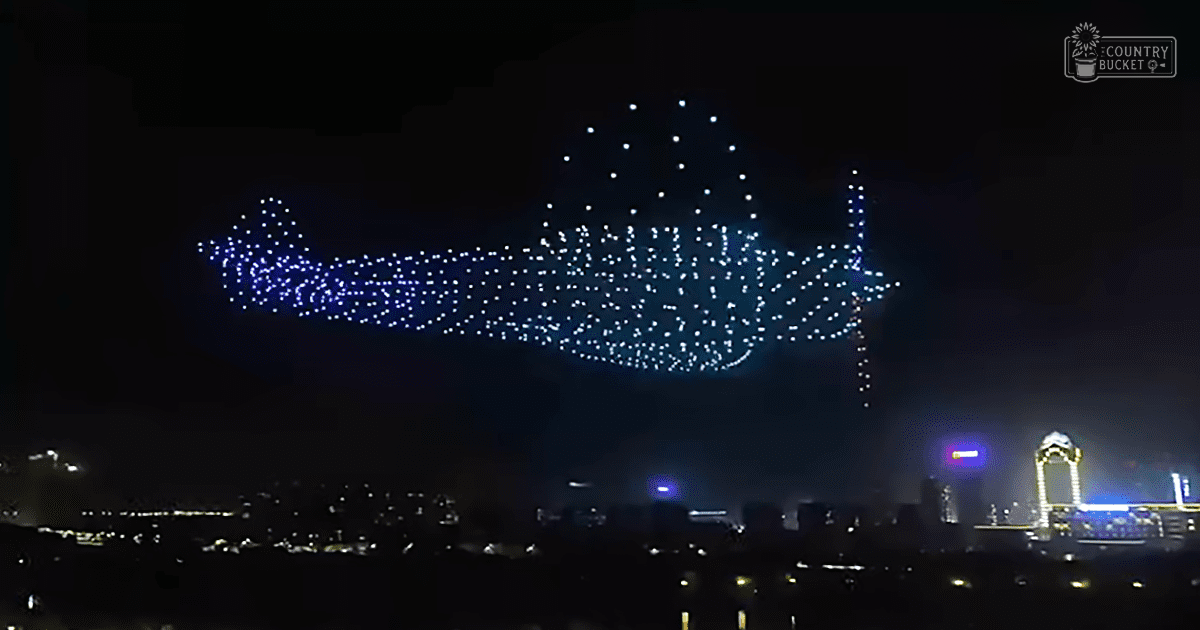An aerial light show forms a large image in the sky over an illuminated cityscape at night with dark blues and blacks contrasted by bright white dots.