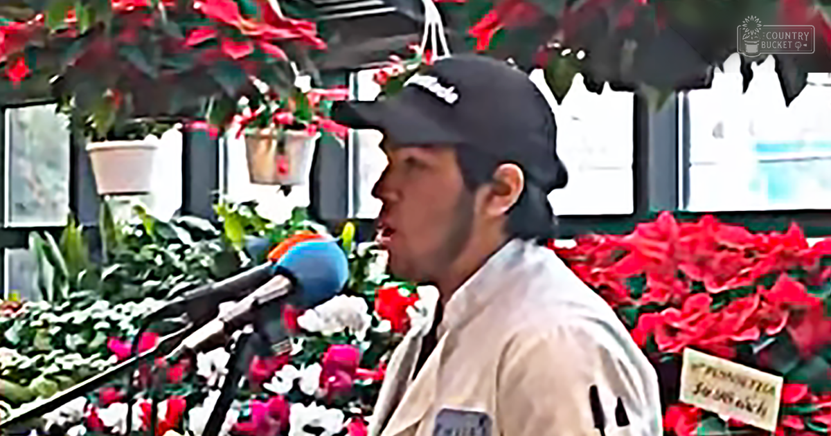 A young adult male singer wearing a light beige jacket and baseball cap, holding a microphone in an indoor setting with hanging potted plants and red flowers.